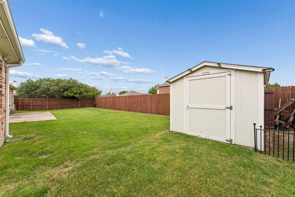 1221 Amistad Drive, Prosper, TX 75078 The property features a spacious backyard with a large lawn, a privacy fence, a concrete patio, and a storage shed