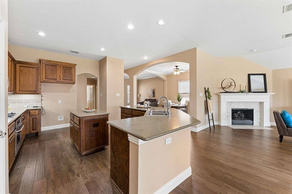 1221 Amistad Drive, Prosper, TX 75078 The property features an open-concept living area with dark wood flooring, a kitchen with rich wood cabinetry and light-colored countertops, and a fireplace with a white mantel