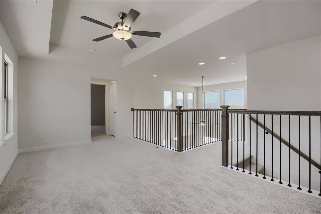 828 Stone Grove Road, Lavon, TX 75166 Carpeted loft area featuring a tray ceiling with a modern ceiling fan, large windows, and a dark wood and iron spindle railing overlooking an open space with additional windows