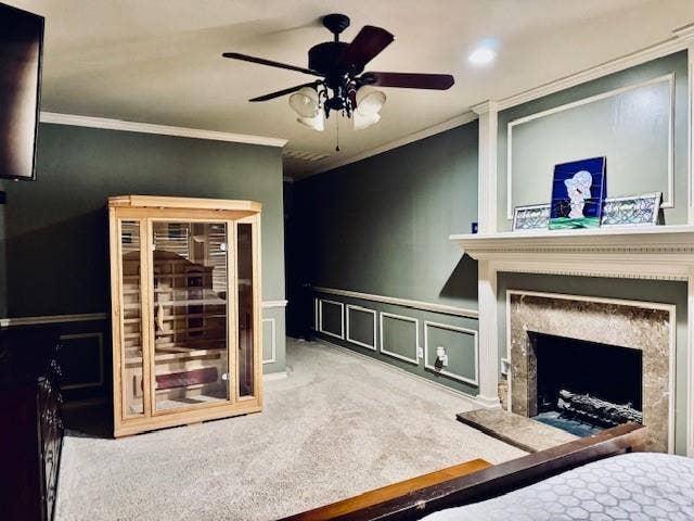 Carpeted bedroom featuring crown molding, a decorative wall, ceiling fan, a fireplace with raised hearth, and a wainscoted wall