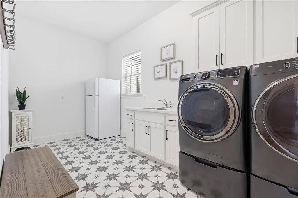 Laundry area featuring patterned floor tile, white shaker cabinetry with black hardware, a utility sink, and a window with blinds