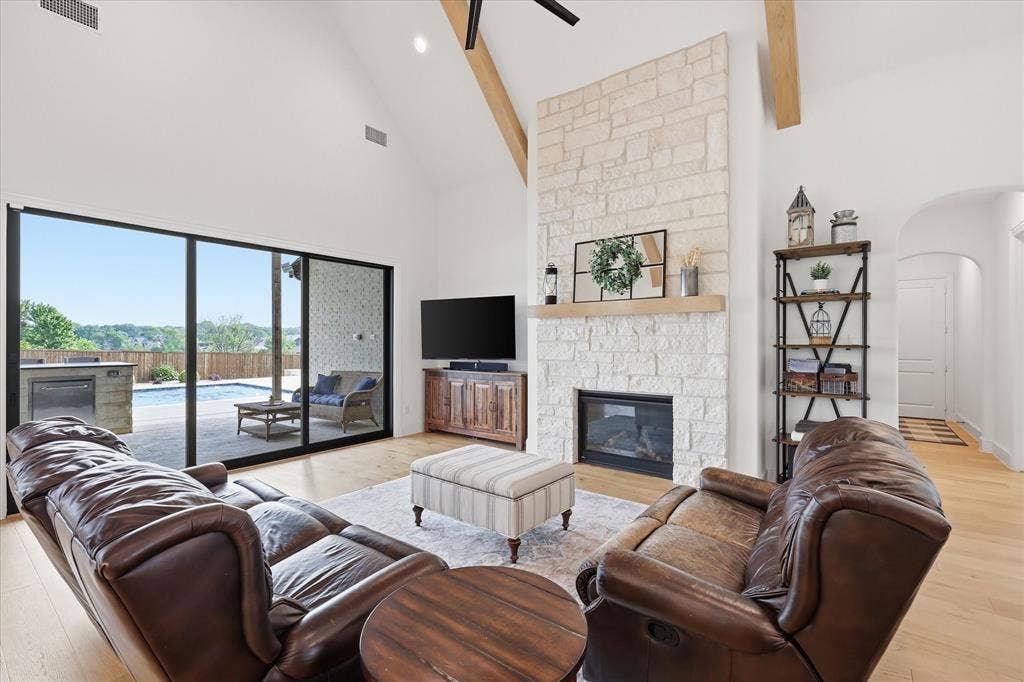 Vaulted ceiling with exposed wood beams, stone fireplace, wood-finish flooring, and expansive sliding glass doors leading to the outdoor space with a built-in grill and swimming pool