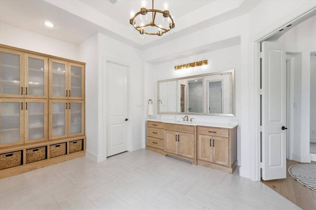 Spacious interior featuring a large wood-finish vanity with a light-colored countertop and double sinks, a substantial built-in storage unit with glass-front cabinetry and base shelving, light-toned tile flooring, and a contemporary chandelier