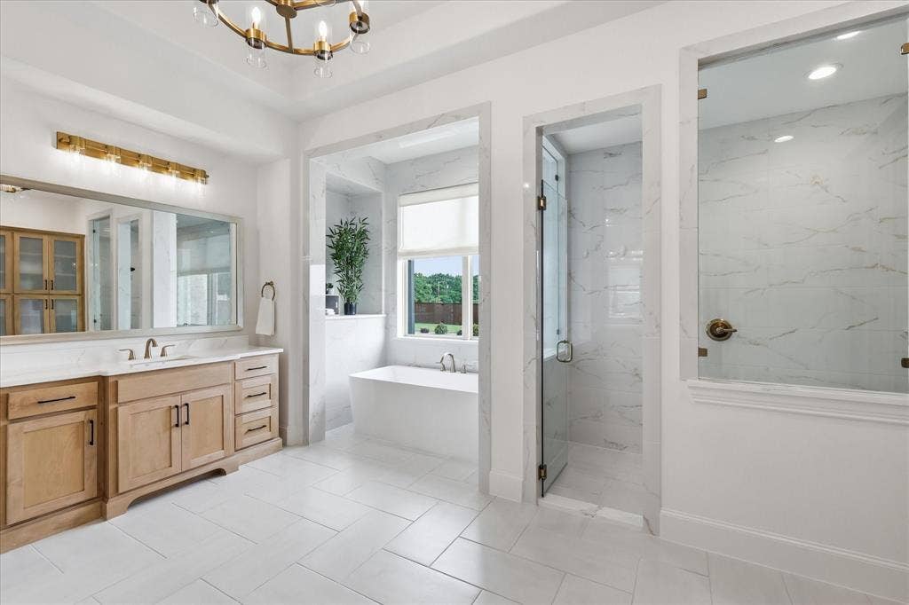 Spacious bathroom featuring a freestanding tub, glass-enclosed shower, light wood-finish vanity with dual sinks, and large-format tile flooring