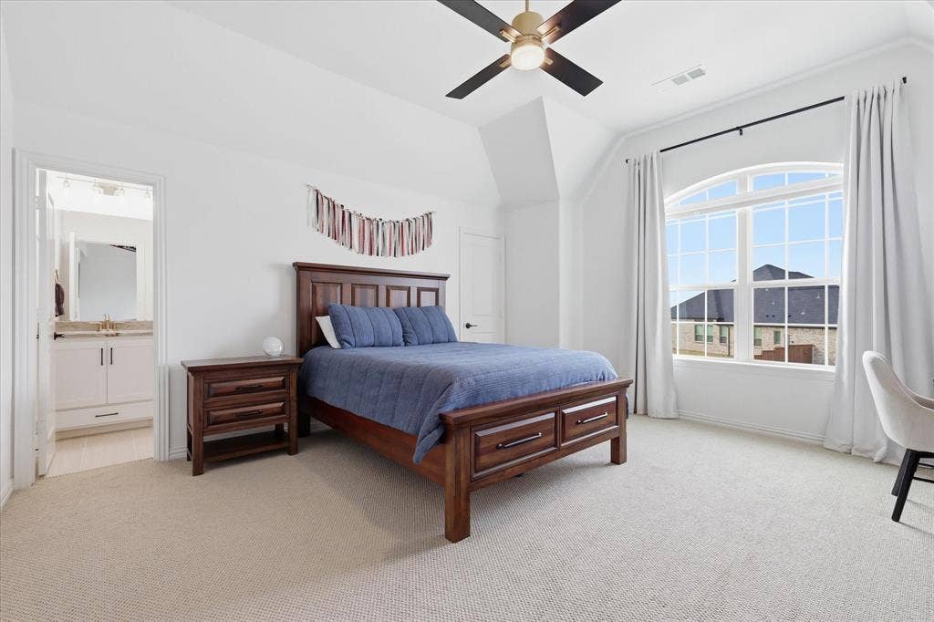 Carpeted bedroom featuring a large arched window, a ceiling fan with integrated lighting, and a white vanity with a gold-toned faucet in the en suite bathroom