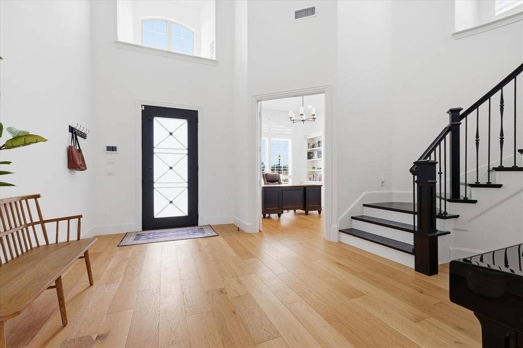 Grand foyer featuring high ceilings, light wood-finish flooring, a black front door with decorative glass panels, and a prominent staircase with black treads and balusters