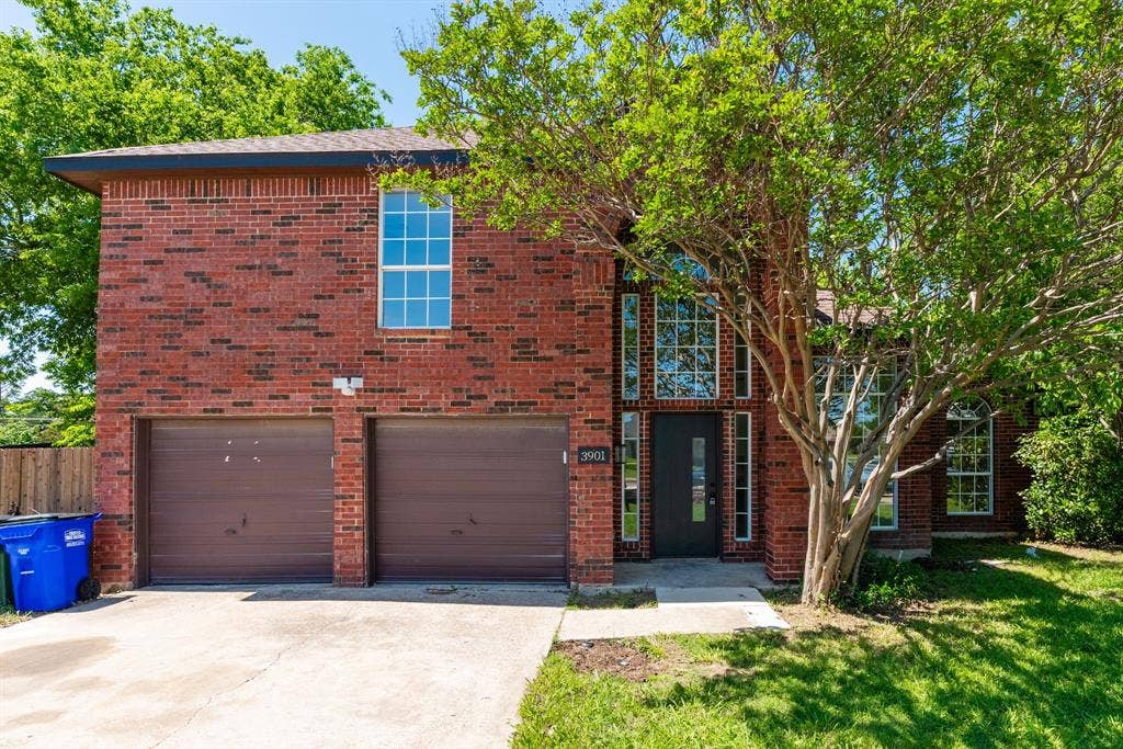Brick exterior residence featuring a two-car garage, mature front yard trees, and a prominent entryway with sidelight windows