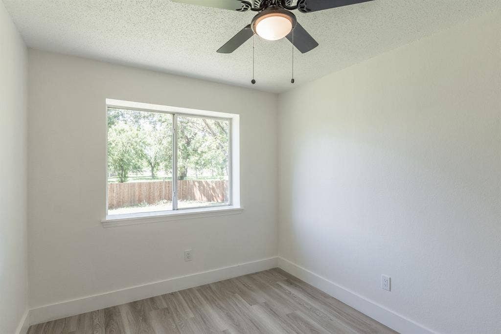 Room featuring a large window, neutral wall paint, wood-finish flooring, white baseboards, and a ceiling fan with integrated lighting