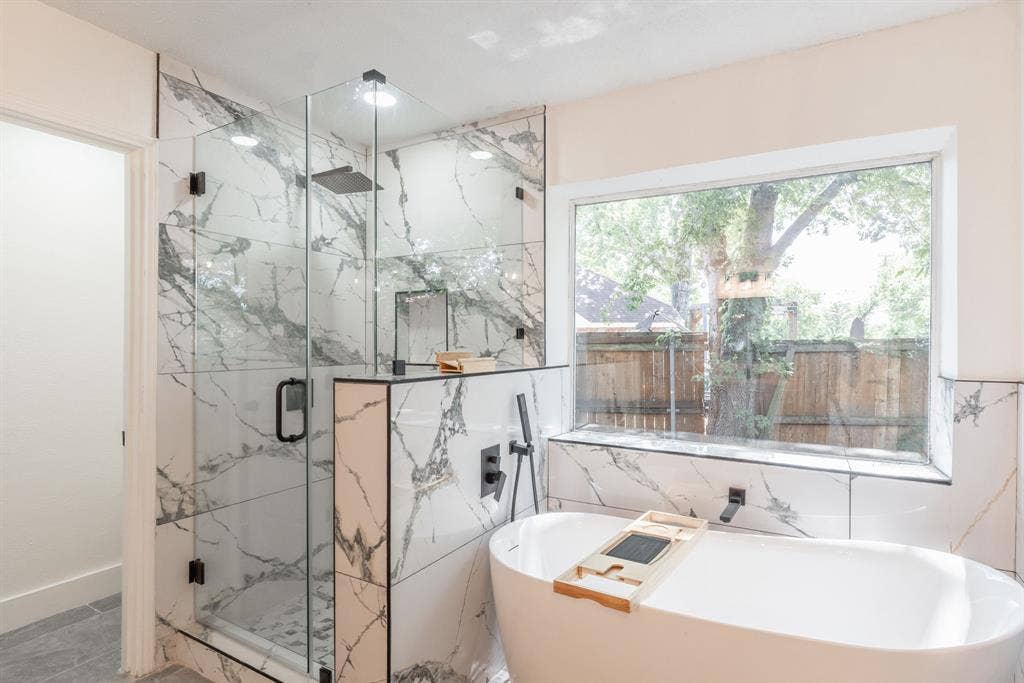 Spacious bathroom featuring a freestanding soaking tub, a large glass-enclosed shower with rain head, marbled tile surround, and expansive window