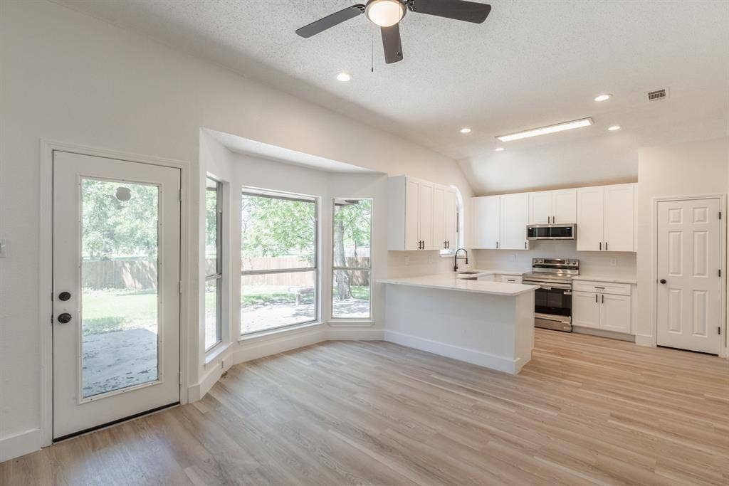 Open-concept interior featuring light wood-finish flooring, a contemporary kitchen with white cabinetry and stainless steel appliances, a multi-pane bay window, recessed lighting, and a ceiling fan