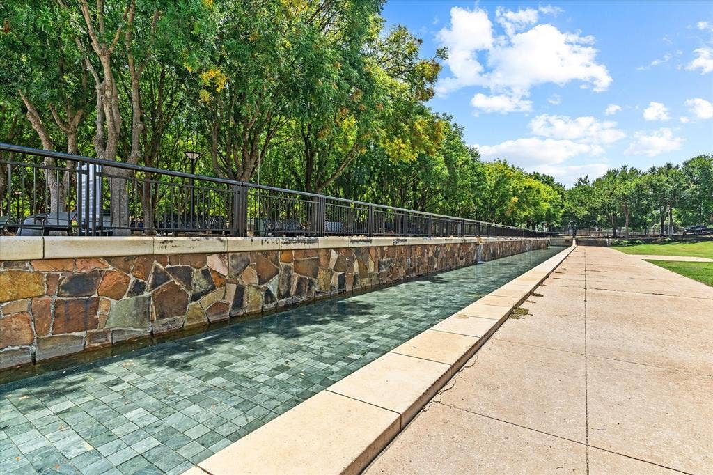 14956 Oak Street, Addison, TX 75001 Decorative water feature with a stone facade, bordered by a concrete pathway and mature trees
