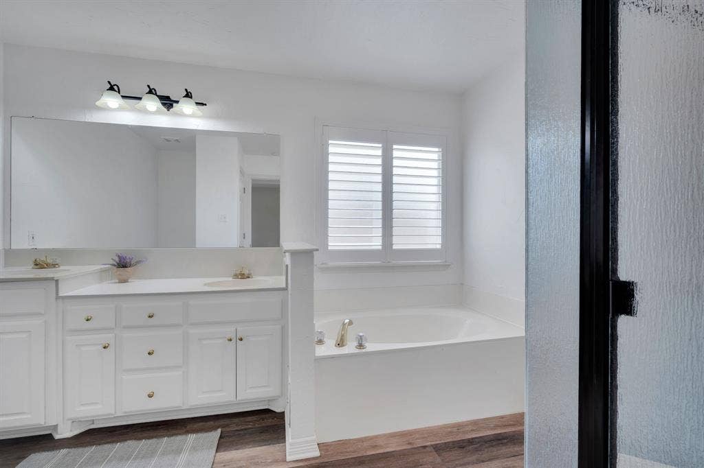 506 Whitney Drive, Midlothian, TX 76065 Bathroom featuring a built-in soaking tub, plantation shutters, white cabinetry with gold hardware, a large vanity mirror, and wood-finish flooring