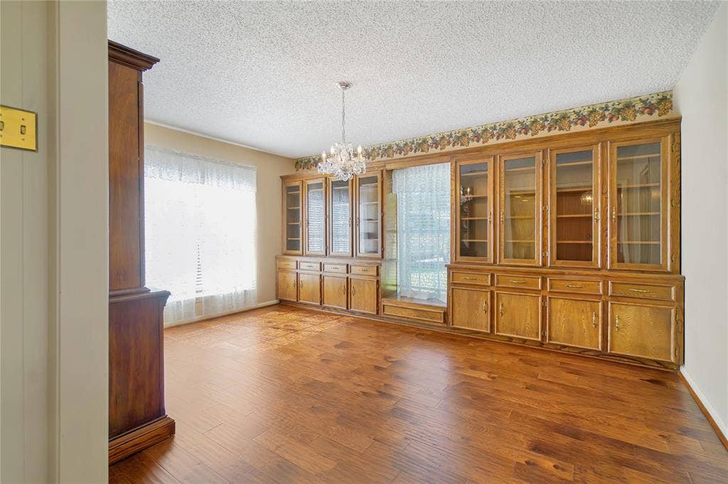 1114 Brookshire Drive, Tyler, TX 75071 Spacious interior room featuring wood-finish flooring, an extensive built-in cabinetry system with glass panel doors, a large window with blinds and sheer curtains, and a prominent chandelier