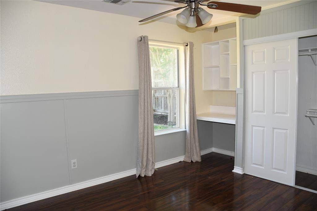Room featuring wood-finish flooring, two-tone walls with wainscoting, a ceiling fan, a built-in desk with shelving, and a sliding closet door