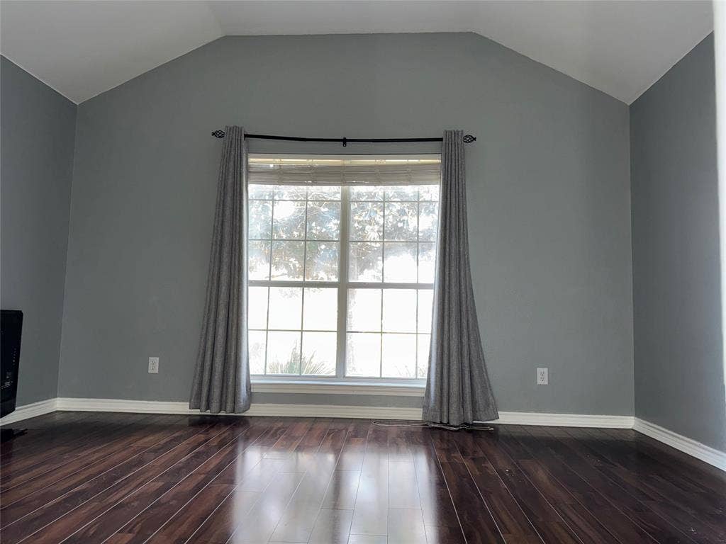 Spacious room featuring vaulted ceilings, a large multi-pane window, dark wood-finish flooring, and light gray wall tones with white trim