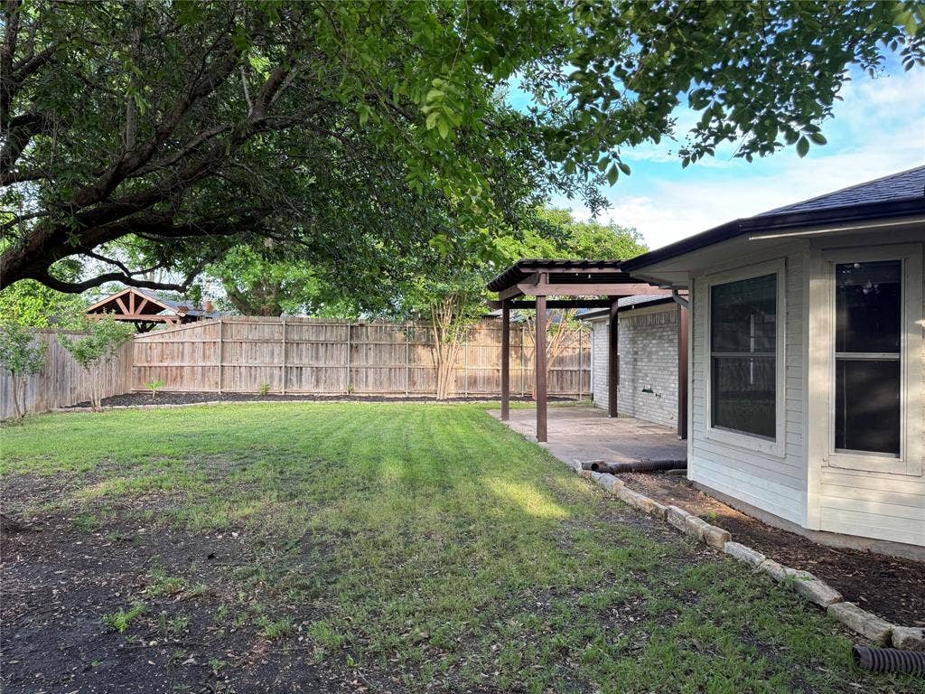 Expansive backyard featuring a lush lawn, mature trees, wood fence, covered patio structure, and light brick exterior
