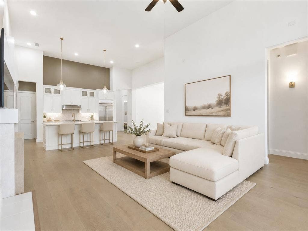 Open-concept living space featuring high ceilings, light wood-finish flooring, and a kitchen with white cabinetry and an island with bar seating. This image has been virtually enhanced for marketing purposes.