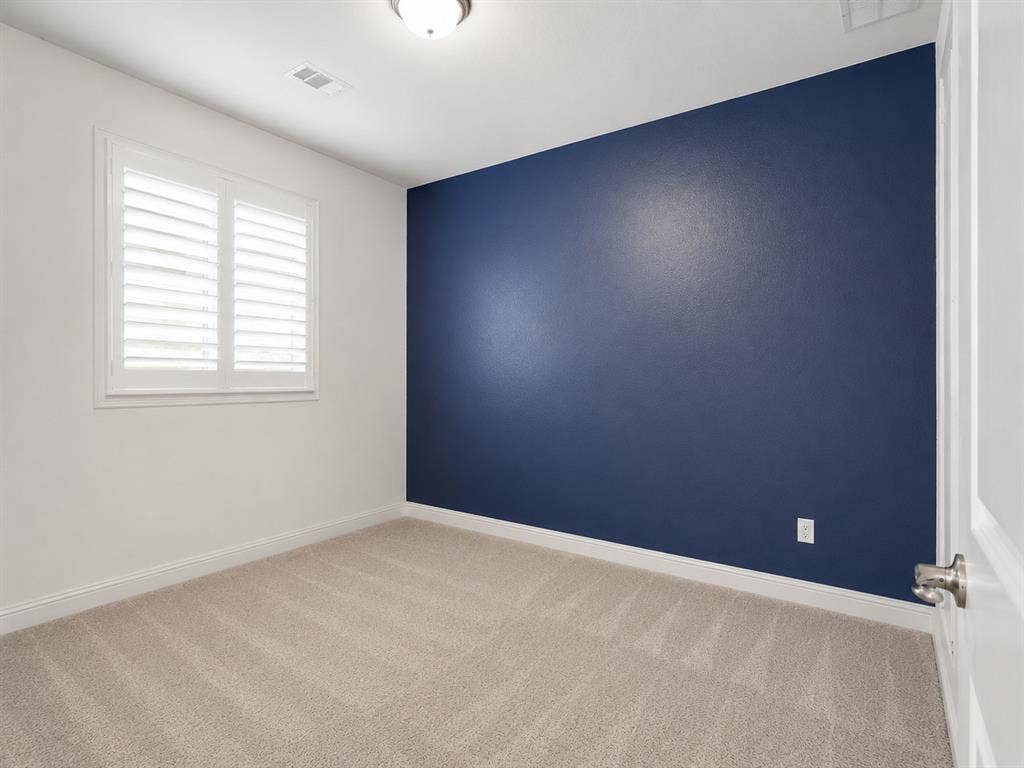 Room featuring a single window with plantation shutters, a blue accent wall, and beige carpeting. This image has been virtually enhanced for marketing purposes.
