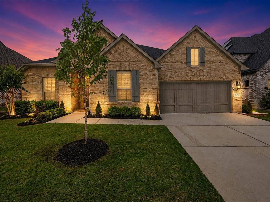 Brick exterior featuring gabled rooflines, decorative shutters, a two-car garage, concrete driveway, and established landscaping with accent lighting