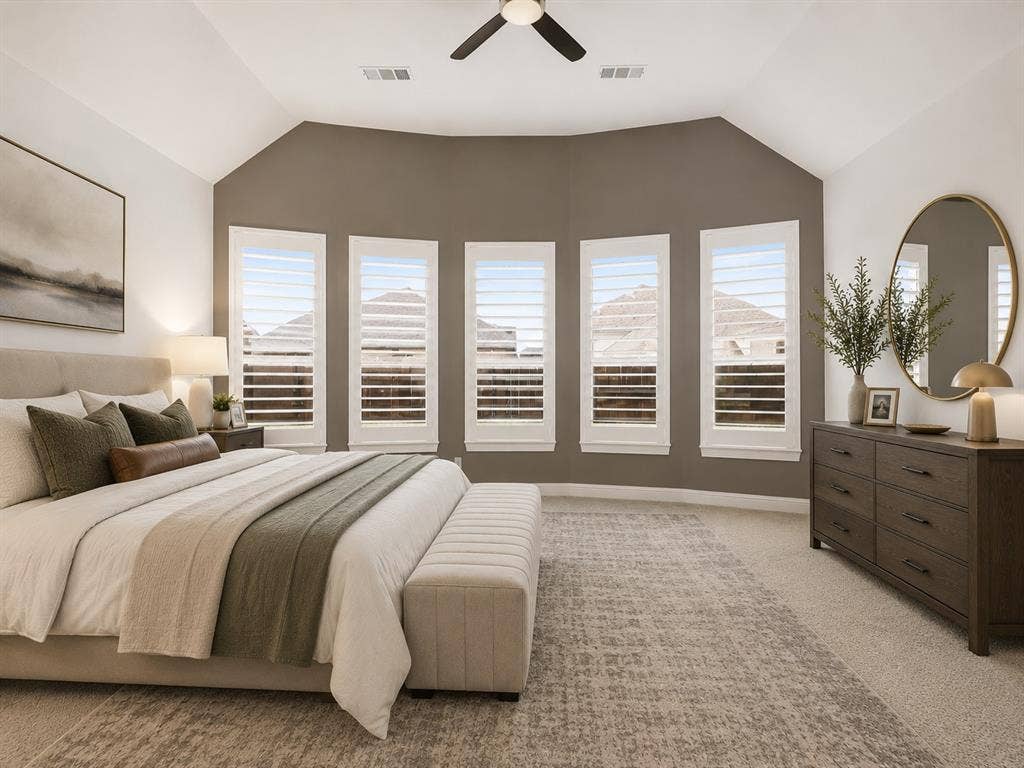 Tray ceiling bedroom featuring a five-panel bay window with white plantation shutters, neutral-toned carpeting, and a ceiling fan with integrated lighting. This image has been virtually enhanced for marketing purposes.