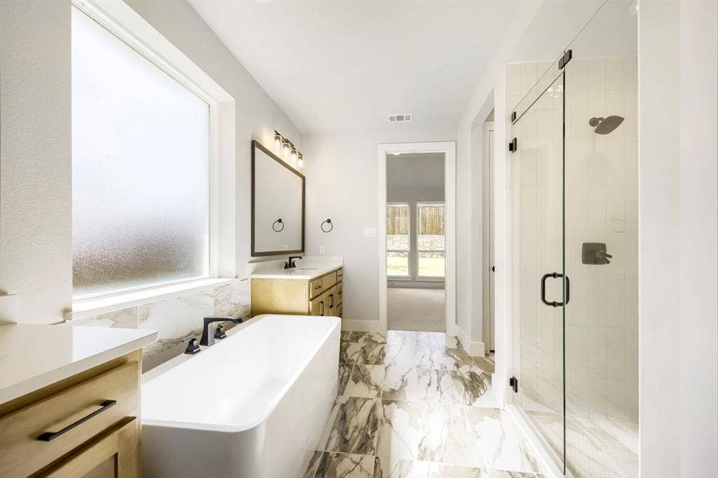 800 Blackberry Road, Haslet, TX 76052 Contemporary bathroom featuring a freestanding soaking tub, a frameless glass shower enclosure, a dual-sink vanity with wood-finish cabinetry, a large rectangular mirror, and marbled tile flooring