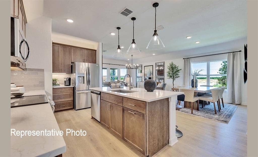 1579 Gentle Night Drive, Forney, TX 75126 Open-concept kitchen featuring an expansive central island with a sink, wood-finish cabinetry, stainless steel appliances, light-toned countertops, and wood-finish flooring