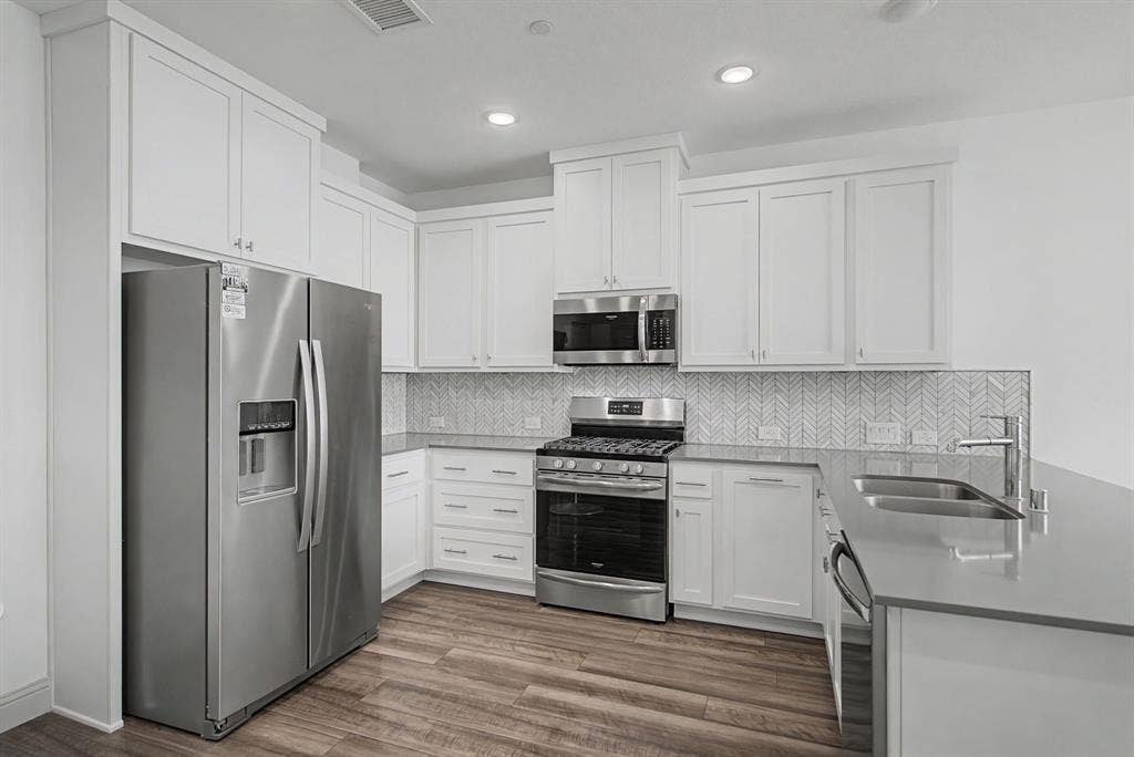 6032 Silverado Trail, McKinney, TX 75070 Kitchen featuring stainless steel appliances, white shaker cabinetry, wood-finish flooring, light-toned countertops, and a herringbone tile backsplash
