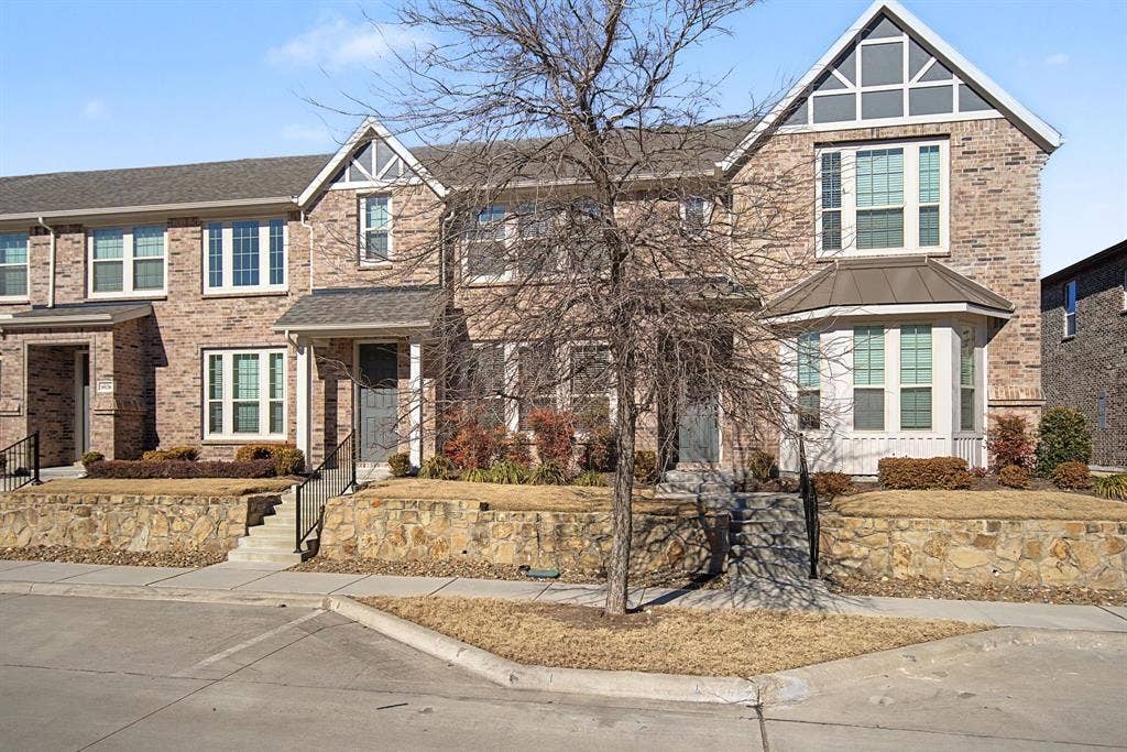 6032 Silverado Trail, McKinney, TX 75070 Brick townhome exterior featuring an arched gable detail, bay window, covered entry, black metal railings, and stone retaining walls