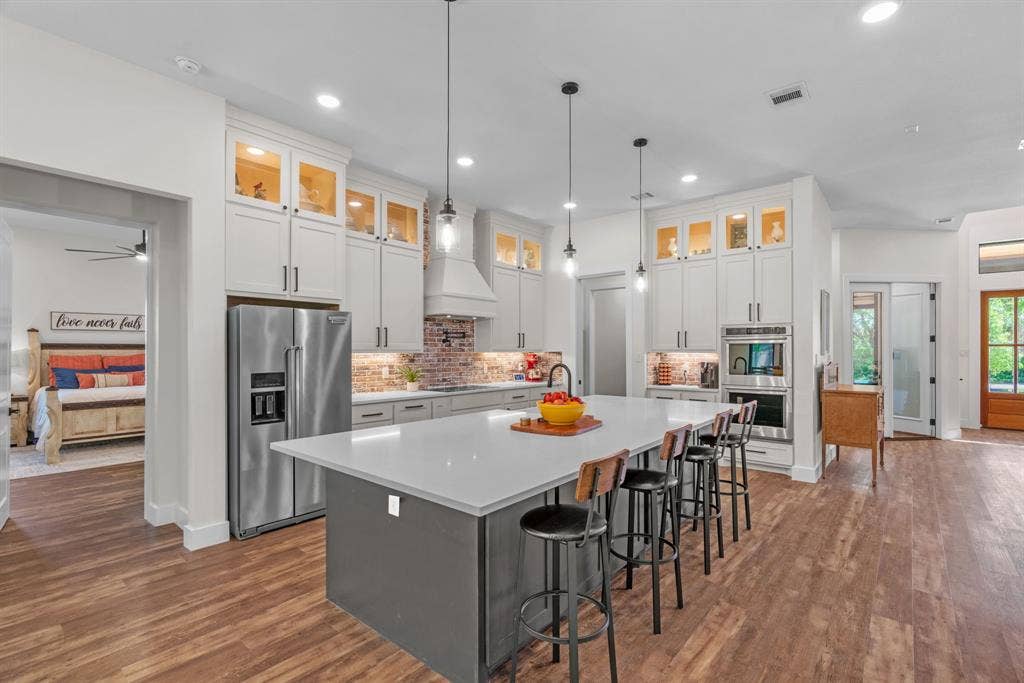 3621 Fritz Lane, Corinth, TX 76208 Gourmet kitchen featuring a large central island with seating, stainless steel appliances, white cabinetry with upper glass display cabinets, brick-finish backsplash, and wood-finish flooring