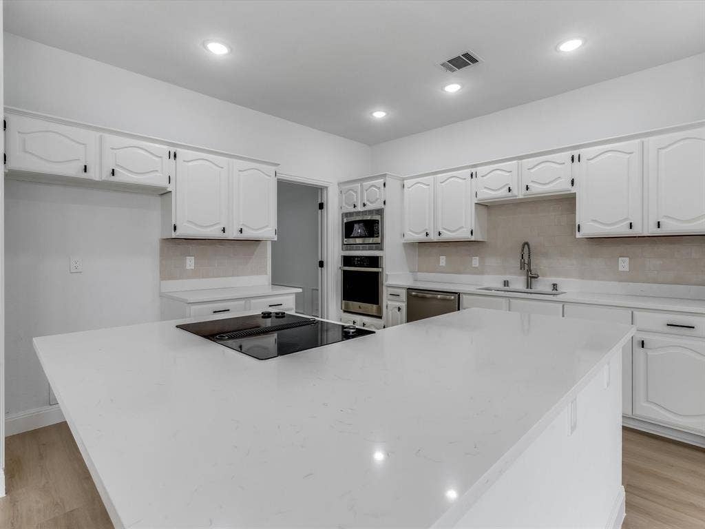 736 Sunkist Lane, Plano, TX 75025 Expansive kitchen island with an integrated cooktop, surrounded by white cabinetry with dark hardware, light-toned backsplash tiling, recessed lighting, and wood-finish flooring