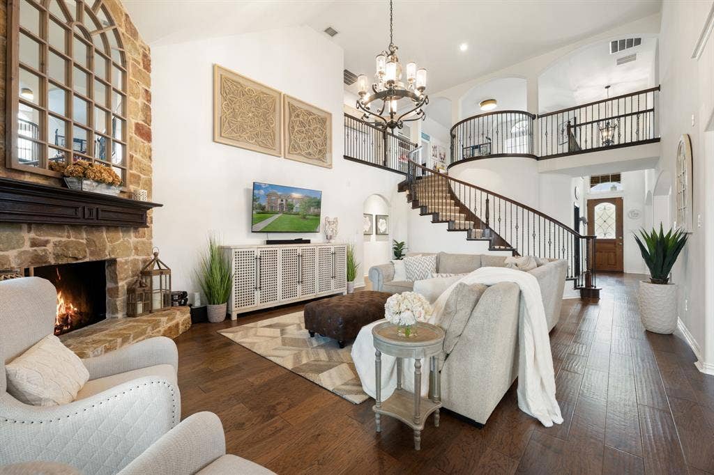 Two-story living area featuring a stone fireplace with a dark wood mantle, wood-finish flooring, a spiral staircase with wrought iron balusters, a second-story balcony, and vaulted ceilings with an ornate chandelier