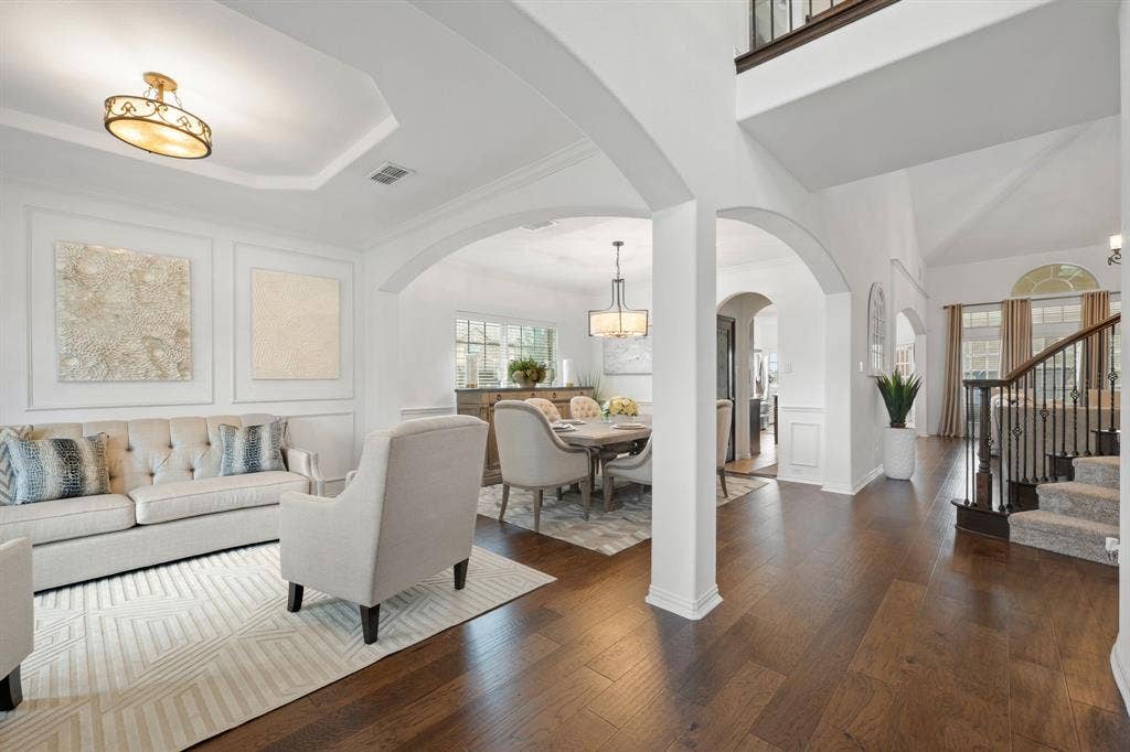 Foyer and living area featuring dark wood-finish flooring, arched doorways, tray ceilings, a grand staircase with wrought iron balusters, and a formal dining room with a chandelier