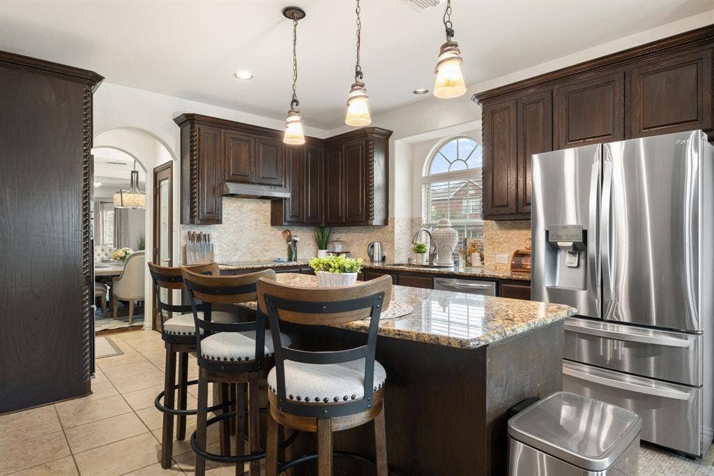 Kitchen featuring dark wood cabinetry, granite countertops, stone tile backsplash, stainless steel appliances, and a central island with pendant lighting