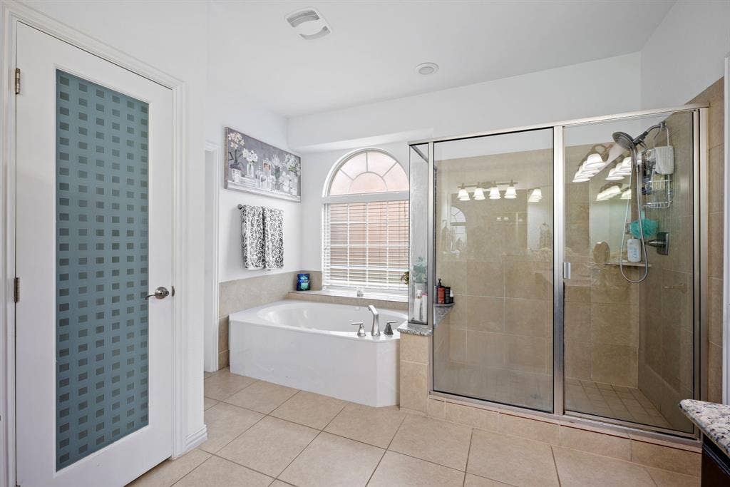 Corner soaking tub with deck-mounted fixtures, separate glass-enclosed shower with tile surround, arched window with horizontal blinds, neutral tile flooring, and a frosted door with a geometric pattern