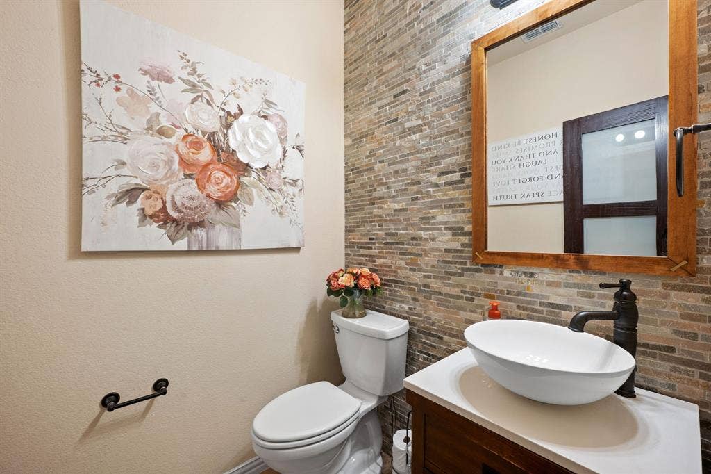 Powder room featuring a stacked stone accent wall, vessel sink with a dark bronze faucet, wood-finish vanity, and a wood-framed mirror