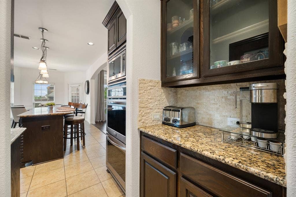 Kitchen featuring granite countertops, stone tile backsplash, and dark wood cabinetry with glass-front uppers