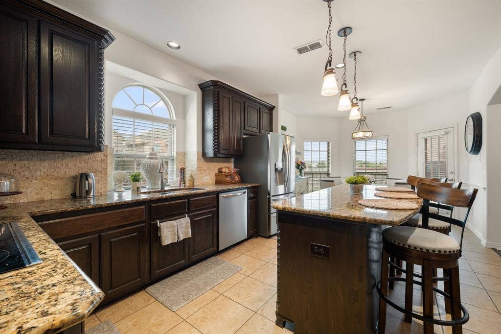 Kitchen featuring dark wood cabinetry, granite countertops, tile flooring, stainless steel appliances, and a central island with pendant lighting