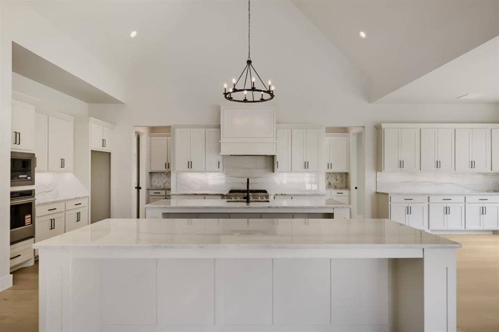794 White Sage Road, Haslet, TX 76052 Expansive kitchen featuring a large central island with a stone countertop, white cabinetry with dark hardware, a built-in range with a custom hood, integrated wall ovens, and vaulted ceilings with recessed lighting and a chandelier