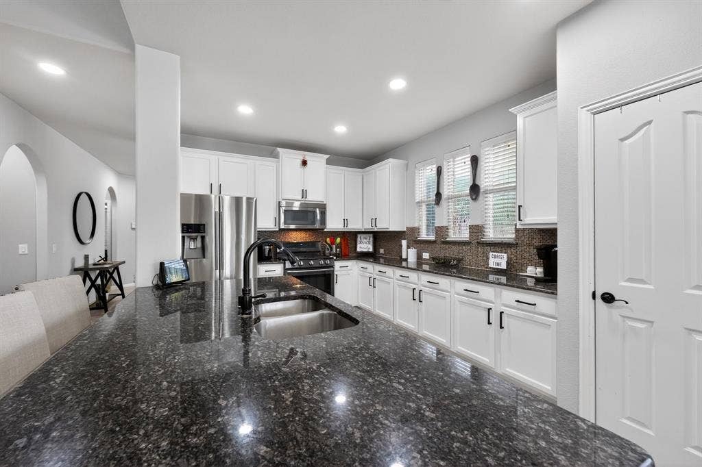2760 Explorador, Grand Prairie, TX 75054 Spacious kitchen featuring a dark granite island, stainless steel appliances, white cabinetry, a large basin sink with a matte black faucet, and a mosaic tile backsplash
