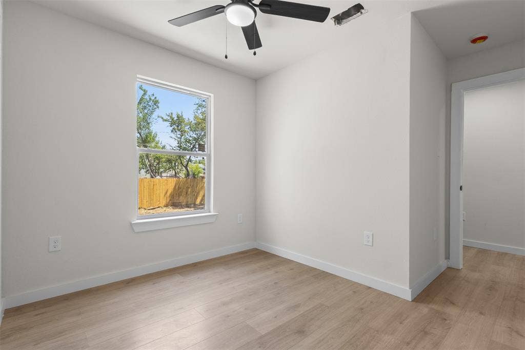 314 NW 15th Street, Grand Prairie, TX 75050 Bright, light-filled bedroom showcasing wood-finish flooring, a large double-hung window with natural landscaping views, ceiling fan with integrated lighting, and clean white walls with electrical outlets