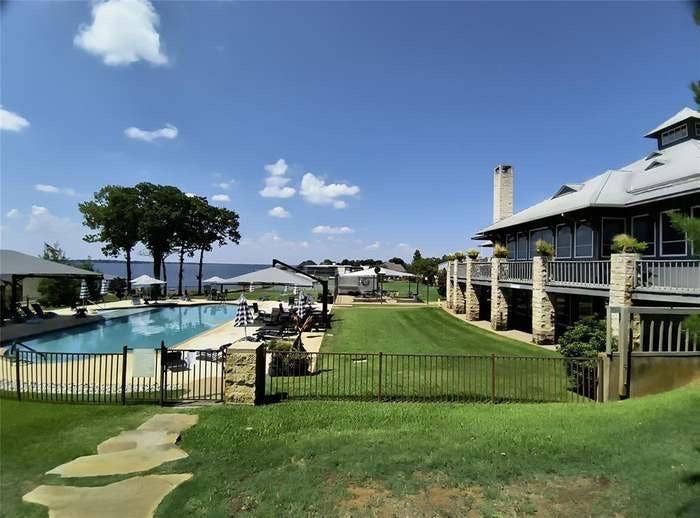 203 Saint Andrews Drive, Mabank, TX 75156 Waterfront property featuring an expansive lawn, a large swimming pool, a second-story balcony with stone pillars, a substantial stone chimney, and mature trees