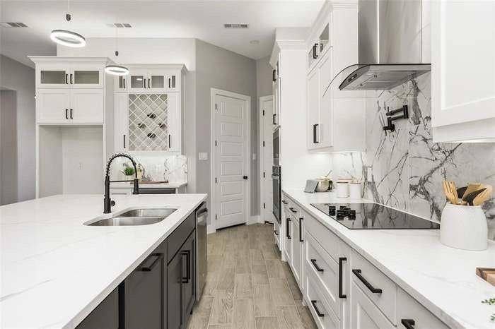 203 Saint Andrews Drive, Mabank, TX 75156 Spacious kitchen featuring an expansive island with an undermount sink and pull-down faucet, white perimeter cabinetry with contrasting hardware, light wood-finish flooring, a built-in wine rack, and a striking natural stone backsplash with a pot filler