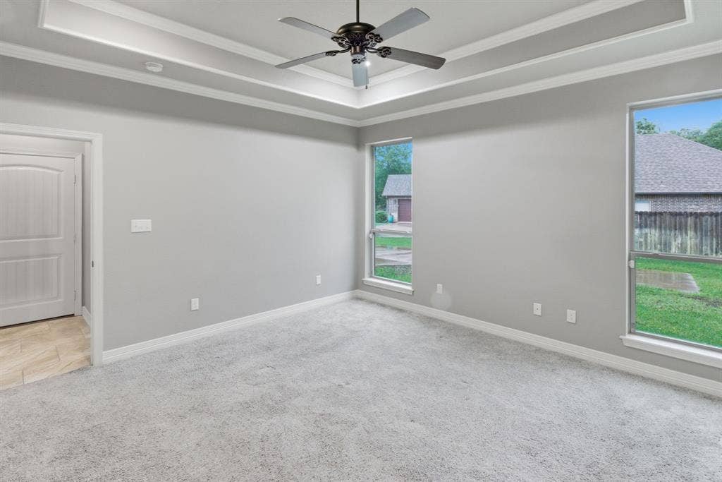 7765 Hickory Spring Lane, Tyler, TX 75703 Spacious room featuring tray ceiling with a ceiling fan, light gray carpeting, two large windows, and a paneled door