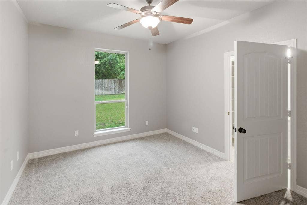 7765 Hickory Spring Lane, Tyler, TX 75703 Neutral-toned room featuring a ceiling fan with integrated lighting, a backyard window overlooking greenery and a wooden fence, plush grey carpeting, and a paneled interior door with matte black hardware