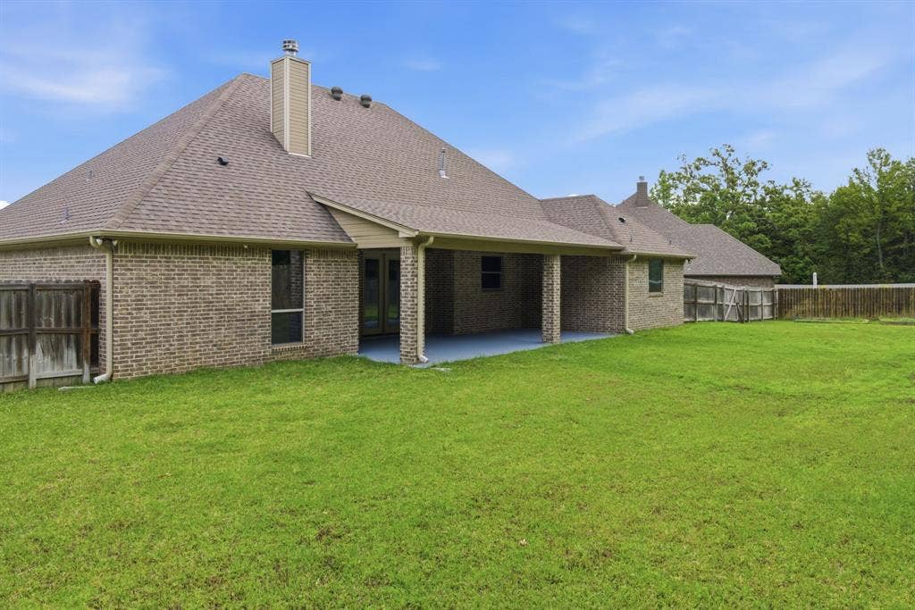 7765 Hickory Spring Lane, Tyler, TX 75703 Expansive grassy yard surrounding a brick property featuring a covered patio with a blue-hued concrete floor, multiple windows, a shingled roof, and a chimney