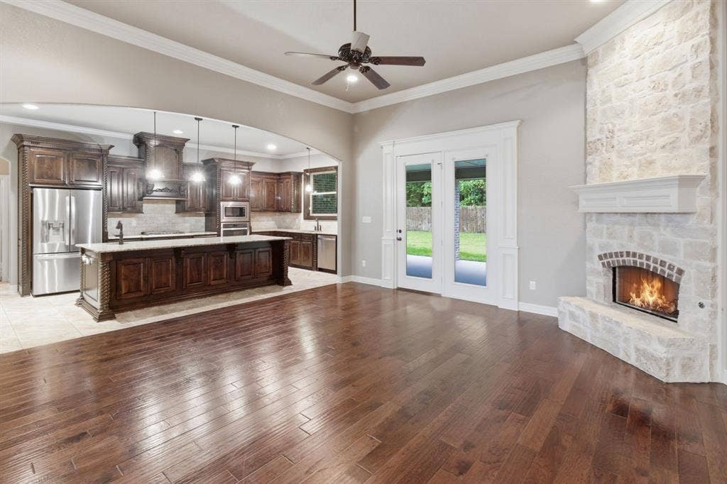7765 Hickory Spring Lane, Tyler, TX 75703 Open-concept living area featuring rich wood-finish flooring, a stone-clad fireplace with a white mantel, and double glass-paneled doors opening to the exterior