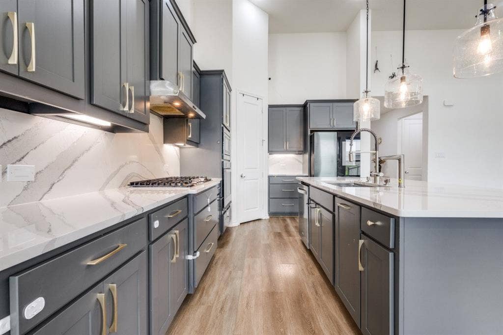 2813 Walker Way, Northlake, TX 76247 Open-concept kitchen featuring gray shaker cabinetry, gold-toned hardware, white countertops with integrated backsplash, wood-finish flooring, and a central island with a stainless steel sink and modern pendant lighting