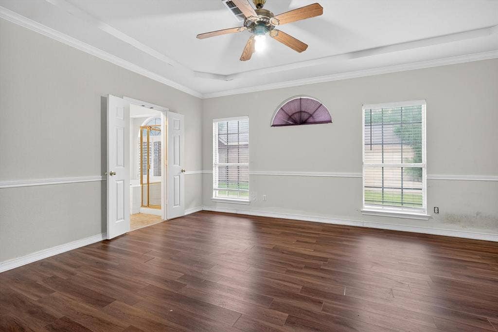 Spacious room featuring rich wood-finish flooring, a tiered tray ceiling with a ceiling fan, and multiple windows including a prominent arched transom window