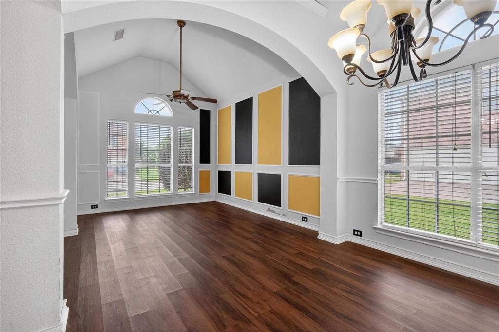 Vaulted ceiling room featuring rich wood-finish flooring, a contemporary accent wall with black and gold panels, and multiple windows with white blinds