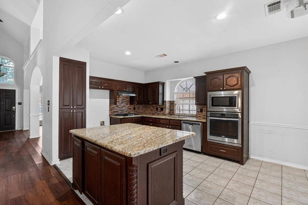 Kitchen featuring a large island with a light-toned countertop, rich wood cabinetry, stainless steel appliances, a tile backsplash, and recessed lighting